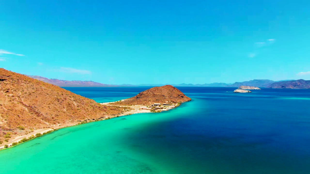 Opening frame over Playa Santispac, Baja California Sur, with turquoise shallows, deeper blue bay water, dry hills at left, distant land on the horizon, and no visible people.
