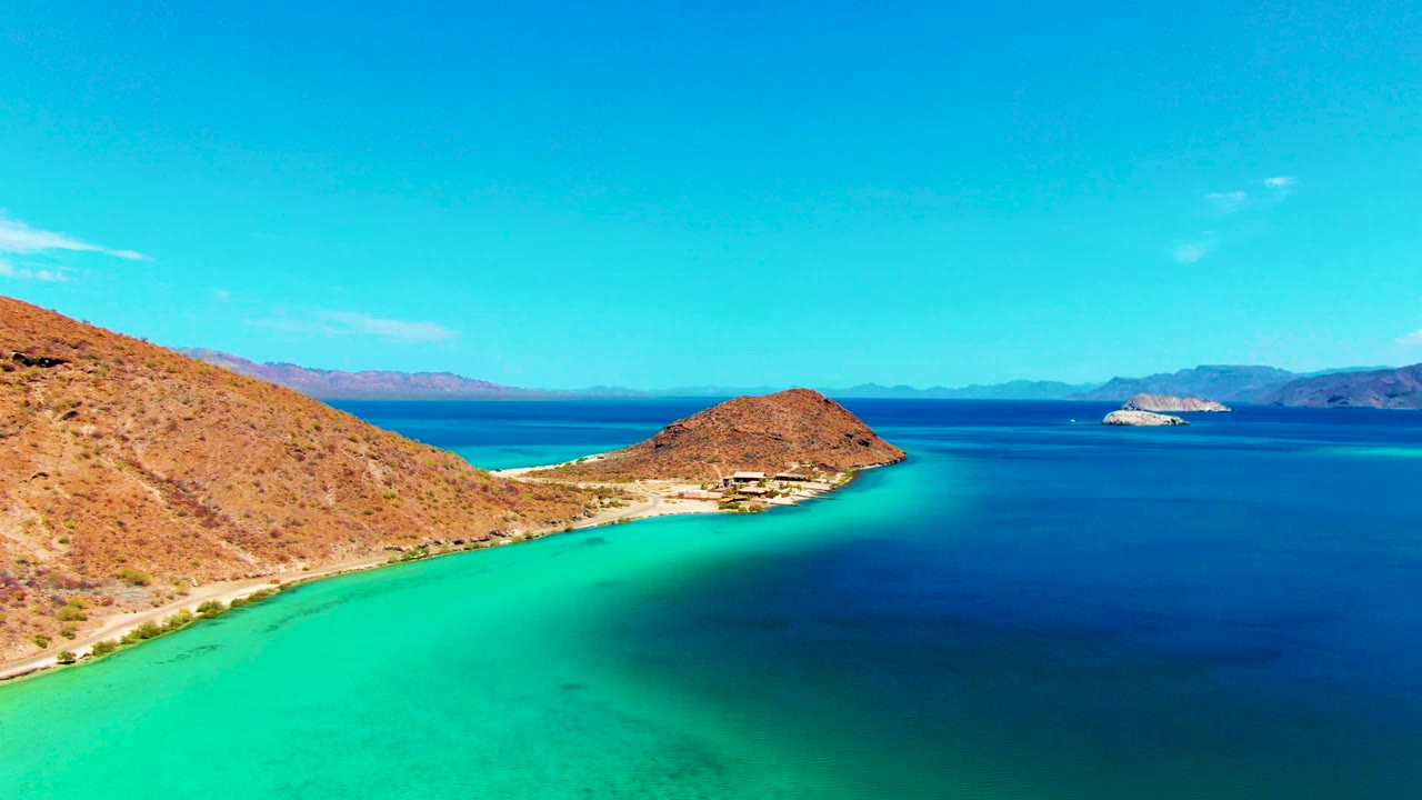 Playa Santispac aerial frame with the hillside filling the left side, a pale beach curve below it, turquoise water in the foreground, and no visible people.