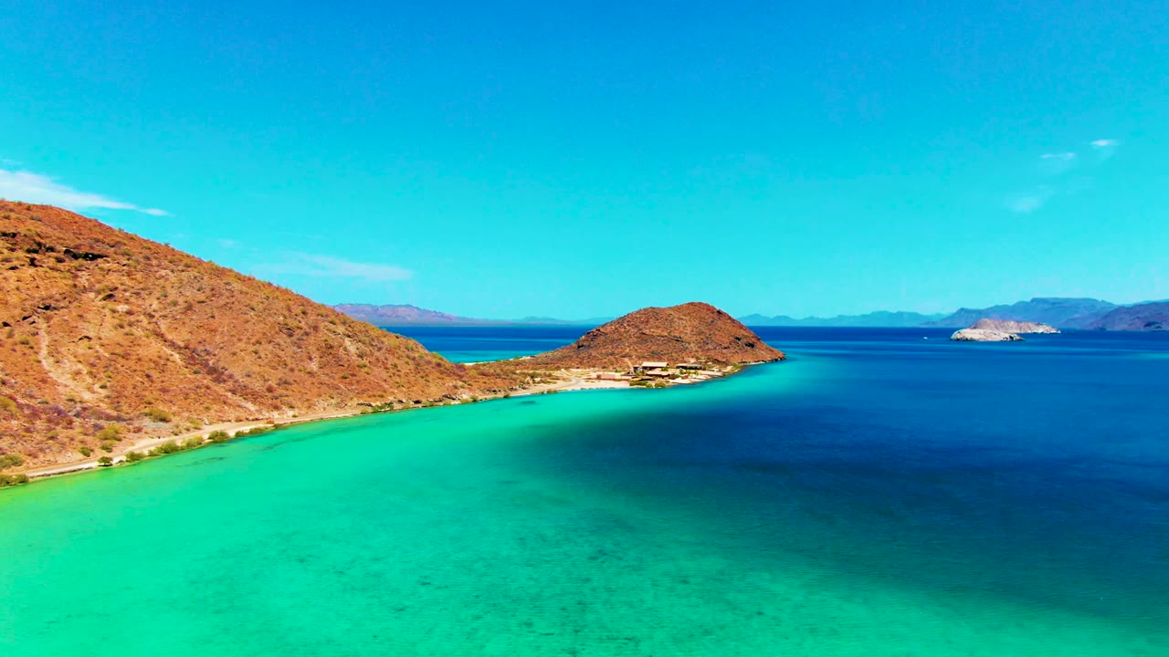 Aerial coastal view with green-blue water, rocky shore, and distant hills.