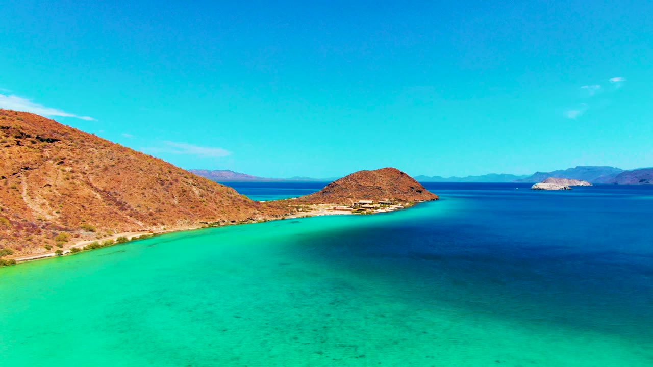 At 0:02, the Playa Santispac coastline curves across the frame, with turquoise water in the foreground, a rocky slope at left, open blue water at right, and no visible people.