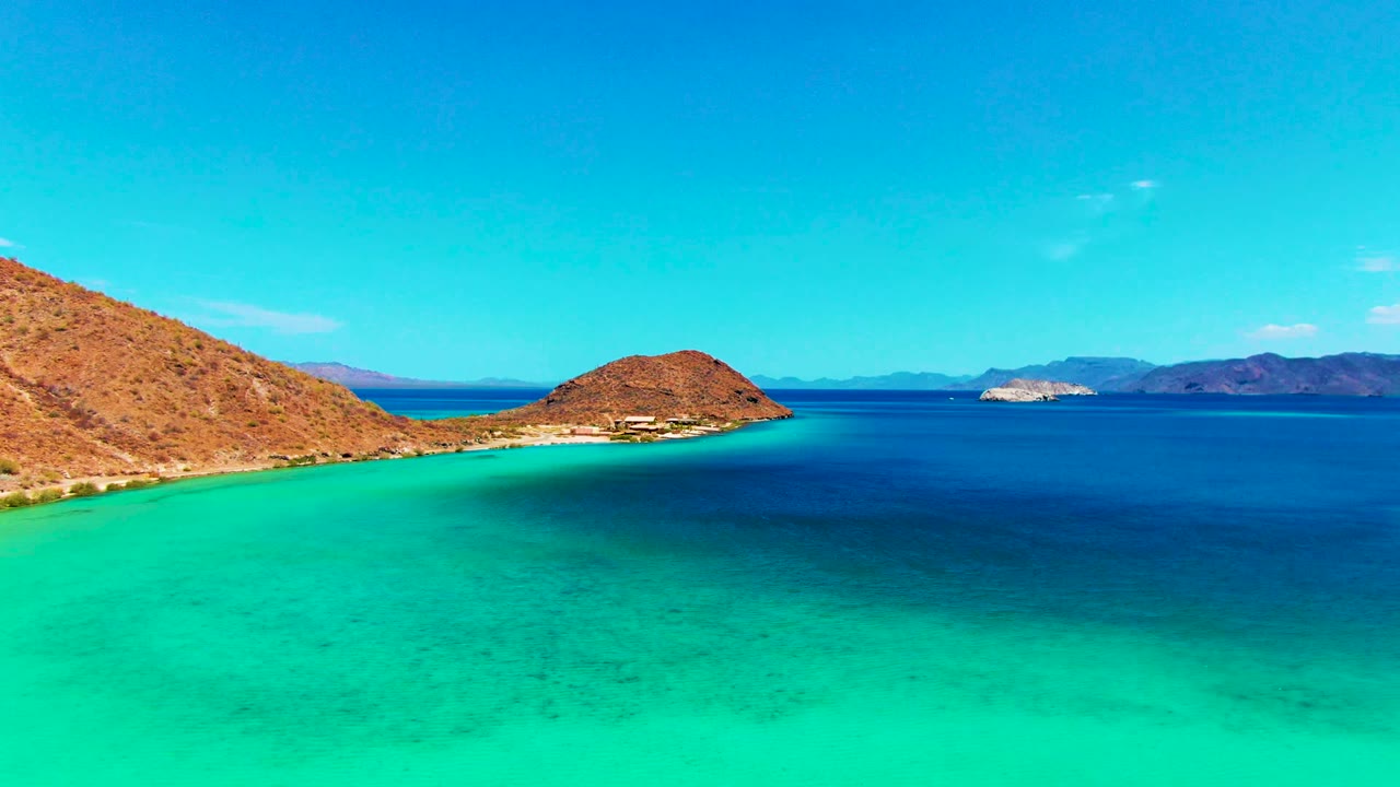 Opening frame at Playa Santispac shows bright turquoise shallow water, a dry rocky slope at left, a small island near center, distant hills, and no visible people.