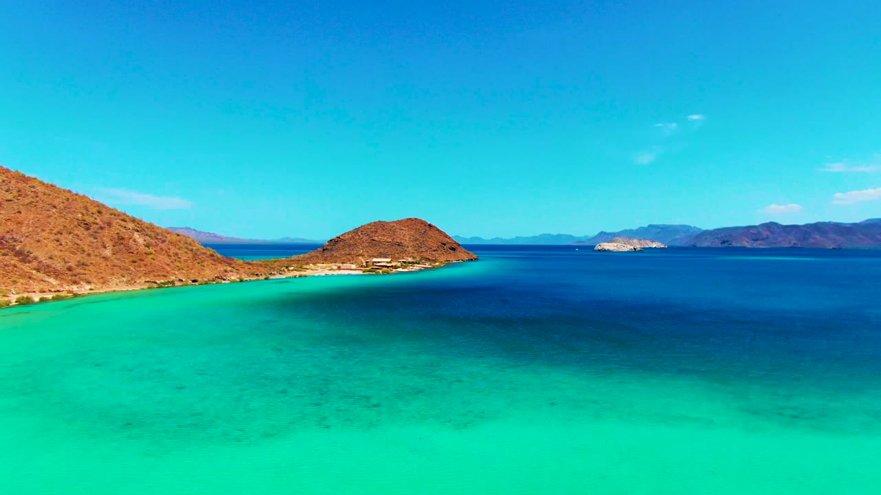 At 0:02, the aerial view shows the same Playa Santispac shoreline with the small island slightly right of center, layered blue water, distant hills, and no visible people.