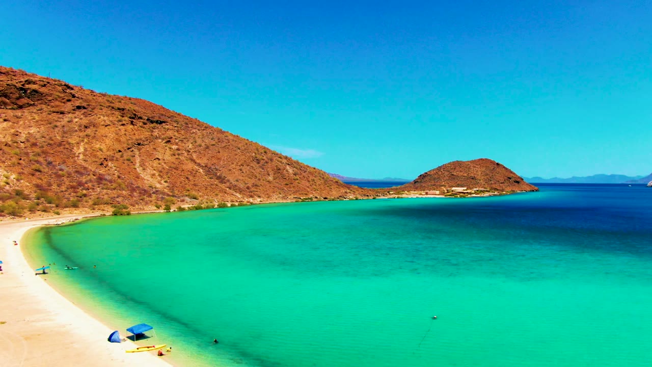 Playa Santispac beach curves beside clear turquoise water, with brown hills in the background and tiny beachgoers near the lower left shore.