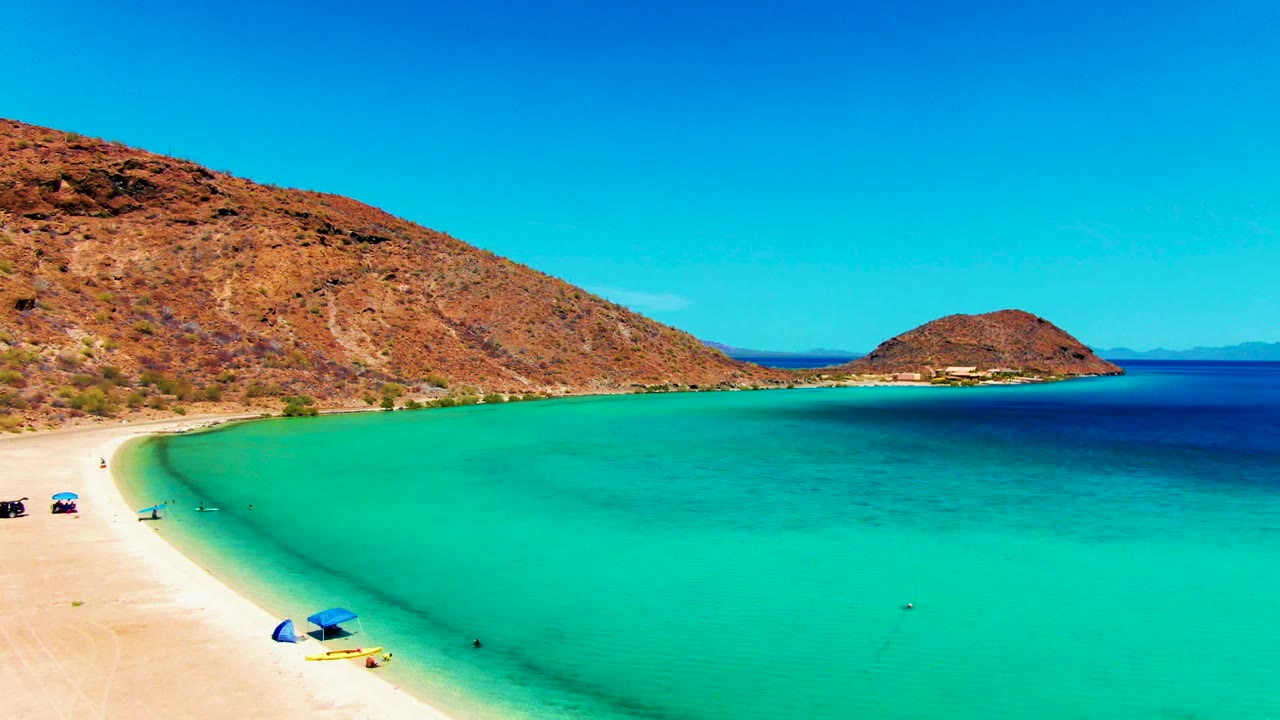 A wider moment at Playa Santispac shows small vehicles and umbrellas on the sand, swimmers as tiny figures, turquoise shallows, and dry hills beyond the bay.