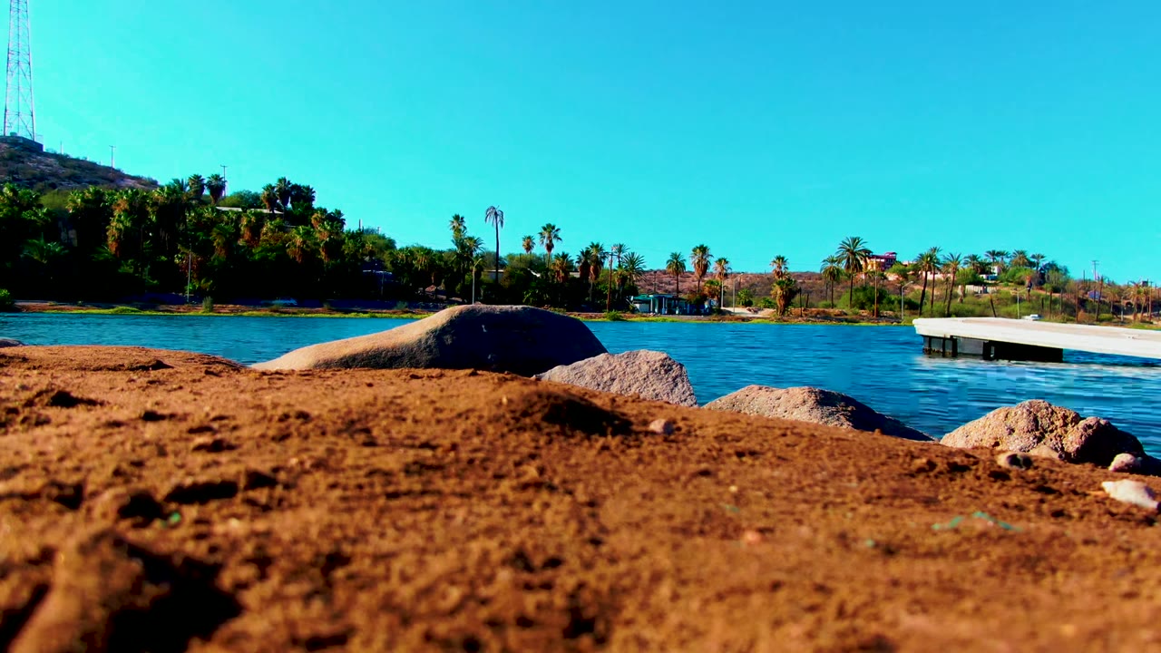 Low-angle Mulege shoreline frame with sand and rocks in the foreground, blue rippled water behind them, a white dock at right, palms and hillside in the distance, and no visible people.