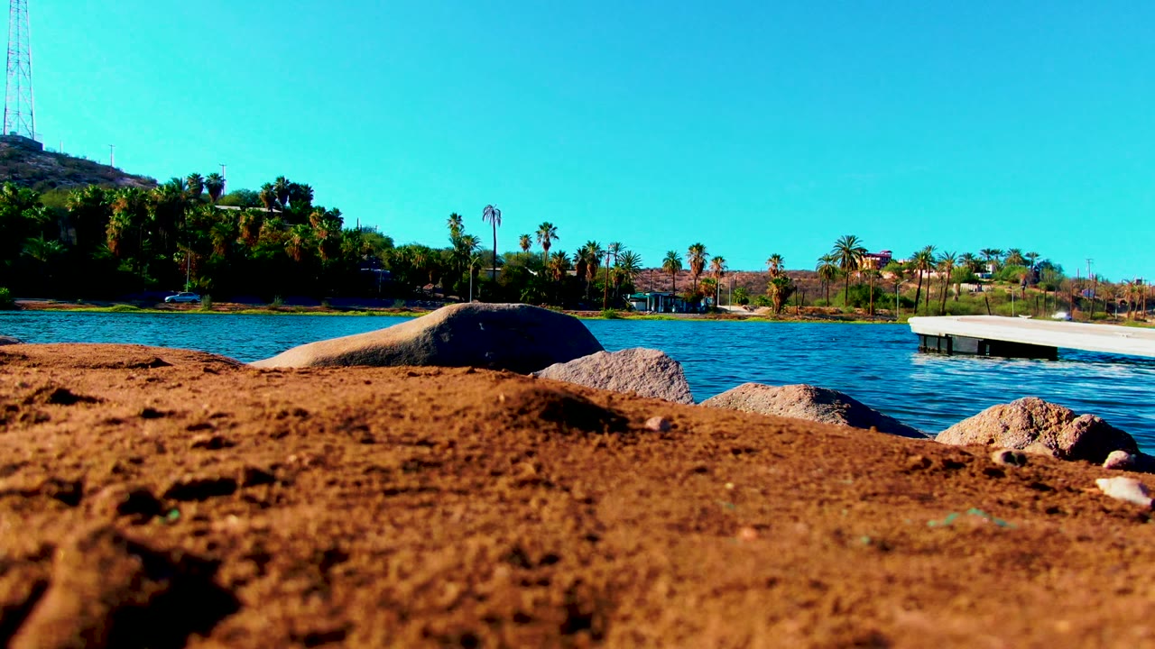 Sandy foreground, boulders, rippled water, palms, and dock under blue sky