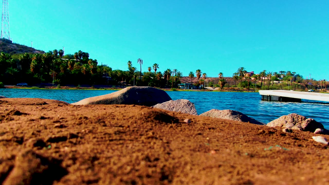 Low Mulege shoreline composition with the sand still filling the bottom of frame, rocks along the edge, blue water in midframe, the dock extending from the right, and no visible people.
