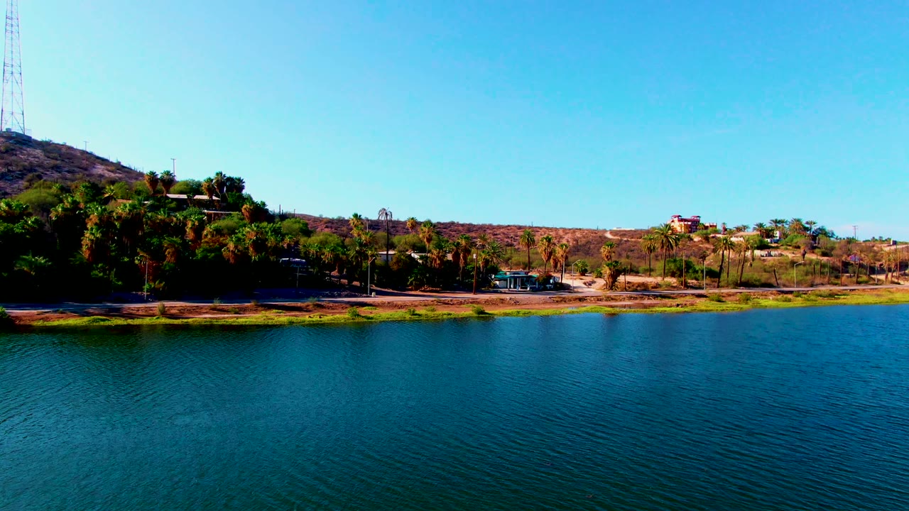 Wide Mulege shoreline frame with turquoise-blue water in front, a strip of green vegetation, palms, hillside buildings, dry ridges, a communications tower, and no visible people.