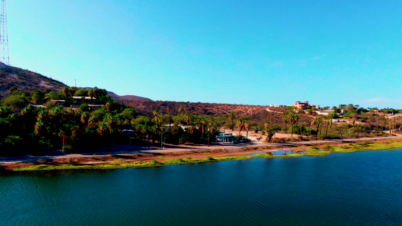 A wide aerial frame over Mulege, Baja California Sur shows blue reservoir water below, a tree-lined shore, dry hillside, scattered buildings, and no visible people.