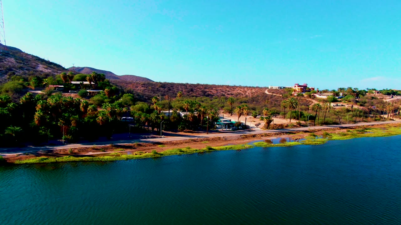 At 0:02, the Mulege aerial frame shows calm reservoir water taking more of the lower view, with palms, scrub-covered hills, scattered structures, and no visible people.