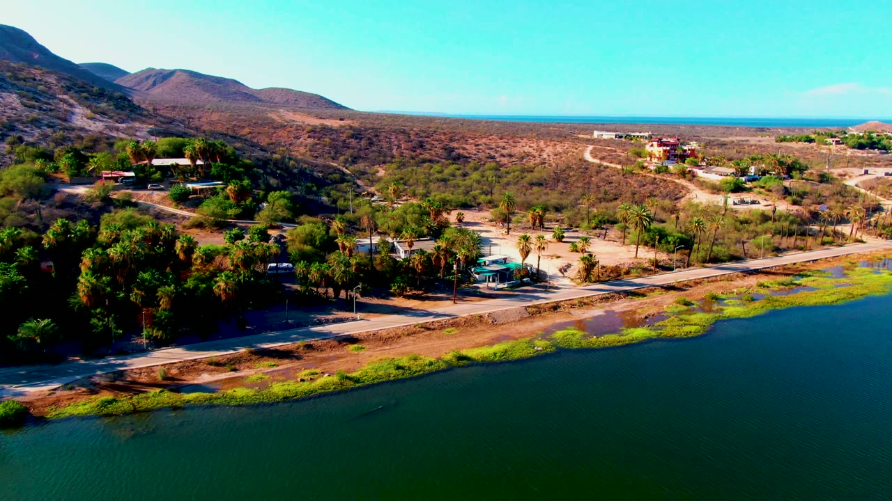 Drone view across water toward a shoreline road, palms, dry valley, hills, and small buildings.