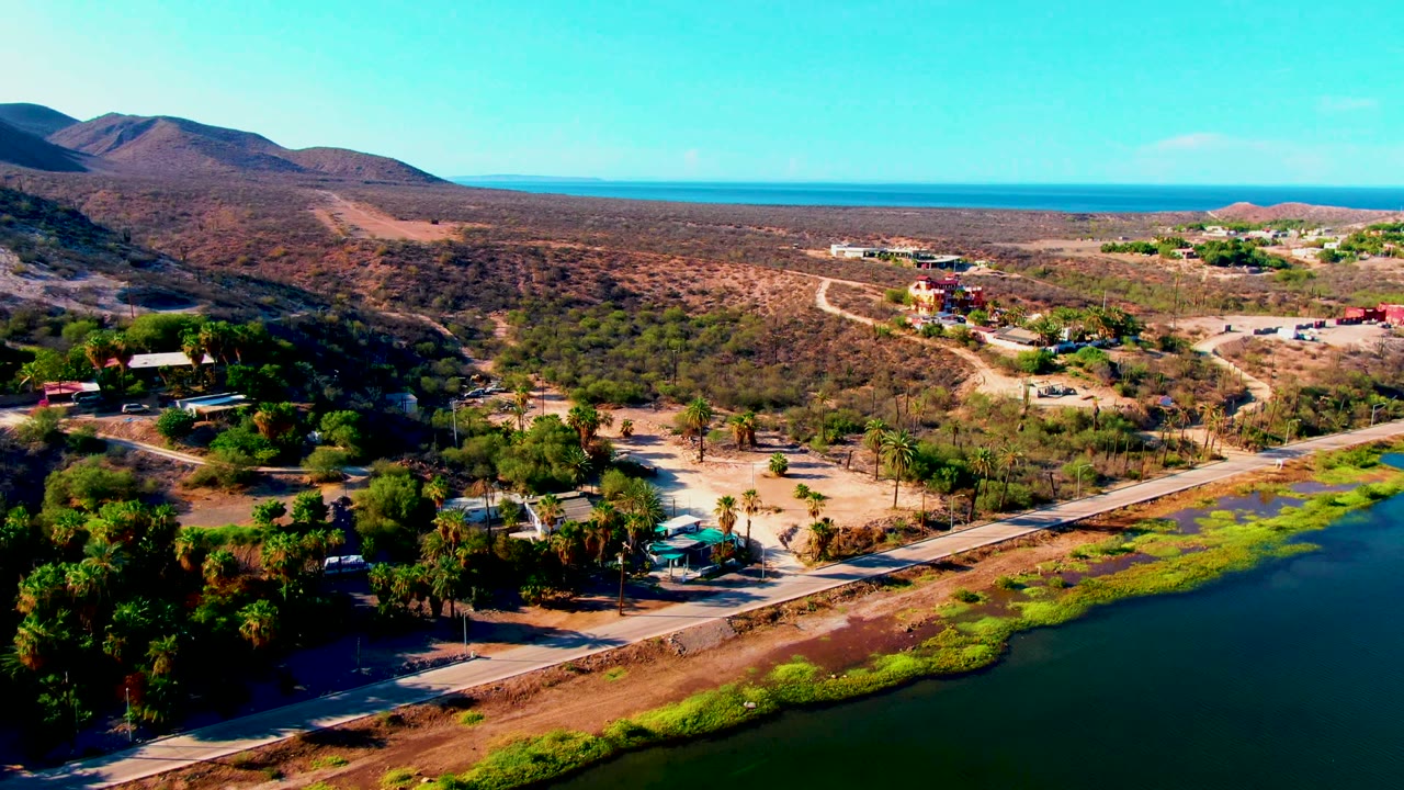 Early aerial frame over Mulege, Baja California Sur, with a roadside waterway in the foreground, dry hills to the left, distant ocean on the horizon, and no visible people.