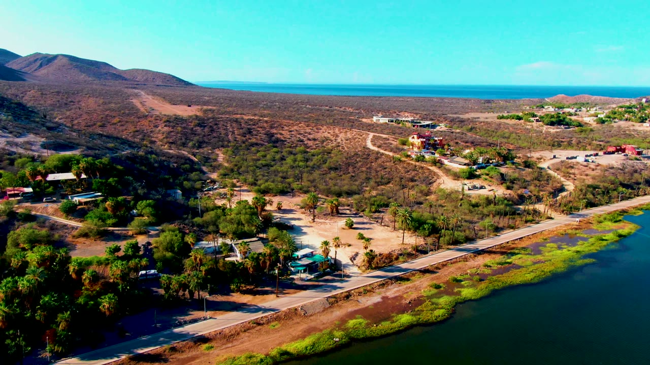 Aerial landscape with road, green shoreline vegetation, dry hills, and distant ocean.
