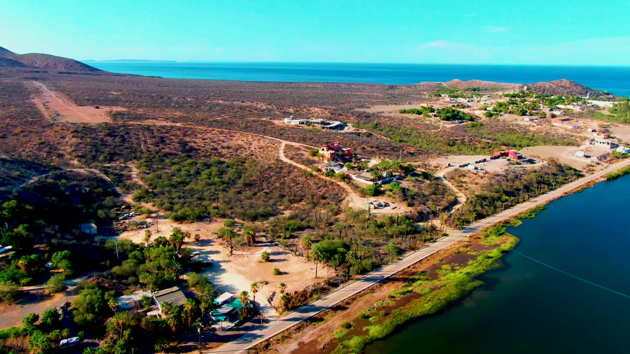 Wide aerial frame over Mulege, Baja California Sur, showing a lagoon-side road, dry scrub hills, scattered buildings, palm trees, and distant ocean, with no visible people.