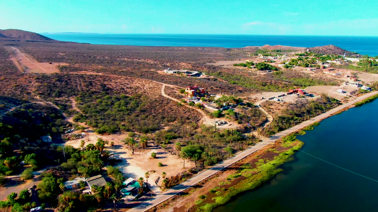 Drone view of lagoon shoreline, road, dry terrain, small buildings, and blue ocean horizon.