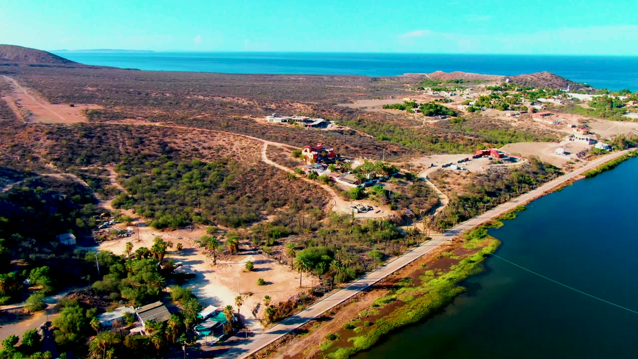 High aerial frame over Mulege showing the road and lagoon edge shifted farther right in the view, with scrubland, a cluster of buildings, low hills, ocean, and no visible people.