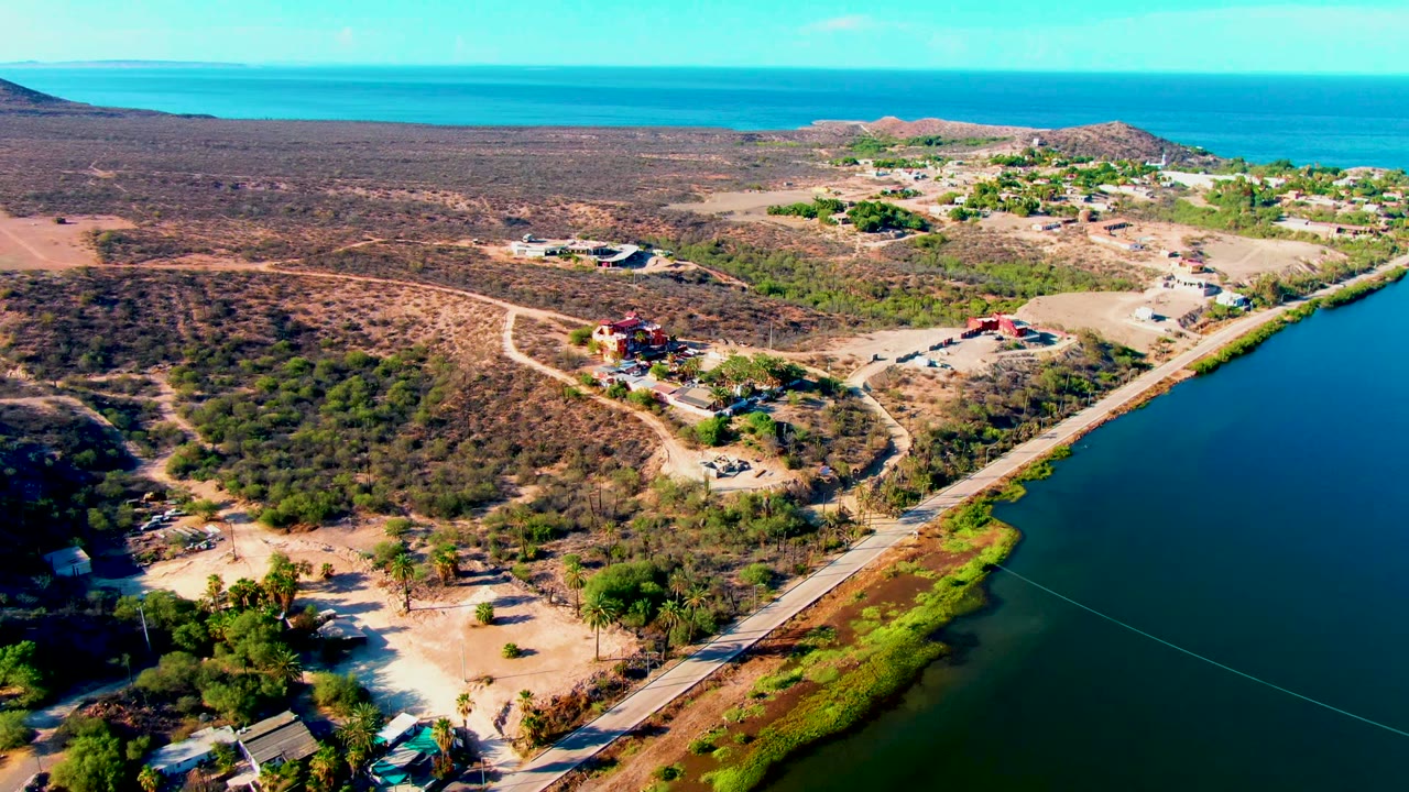 Aerial frame over Mulege, Baja California Sur showing a road along green-blue water, dry brush, scattered buildings, low hills, ocean horizon, and no visible people.