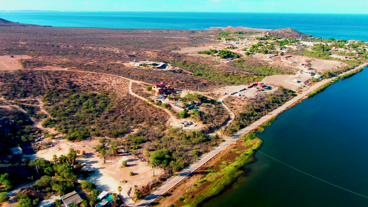 Mulege aerial frame with a paved road tracing the waterline, sparse brown-green vegetation, a few buildings near dirt lots, distant low hills, ocean horizon, and no visible people.