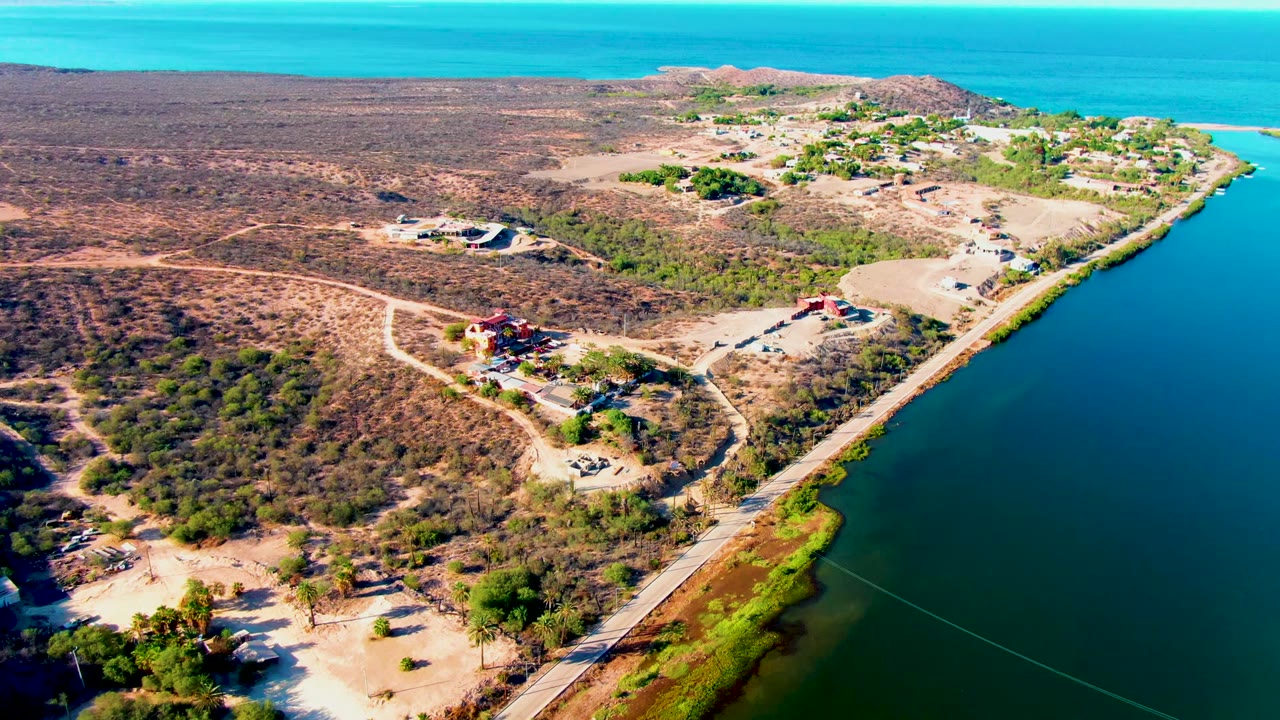 Opening aerial frame over Mulege, Baja California Sur, showing a coastal road along blue-green water, dry scrubland, scattered buildings, and no visible people.