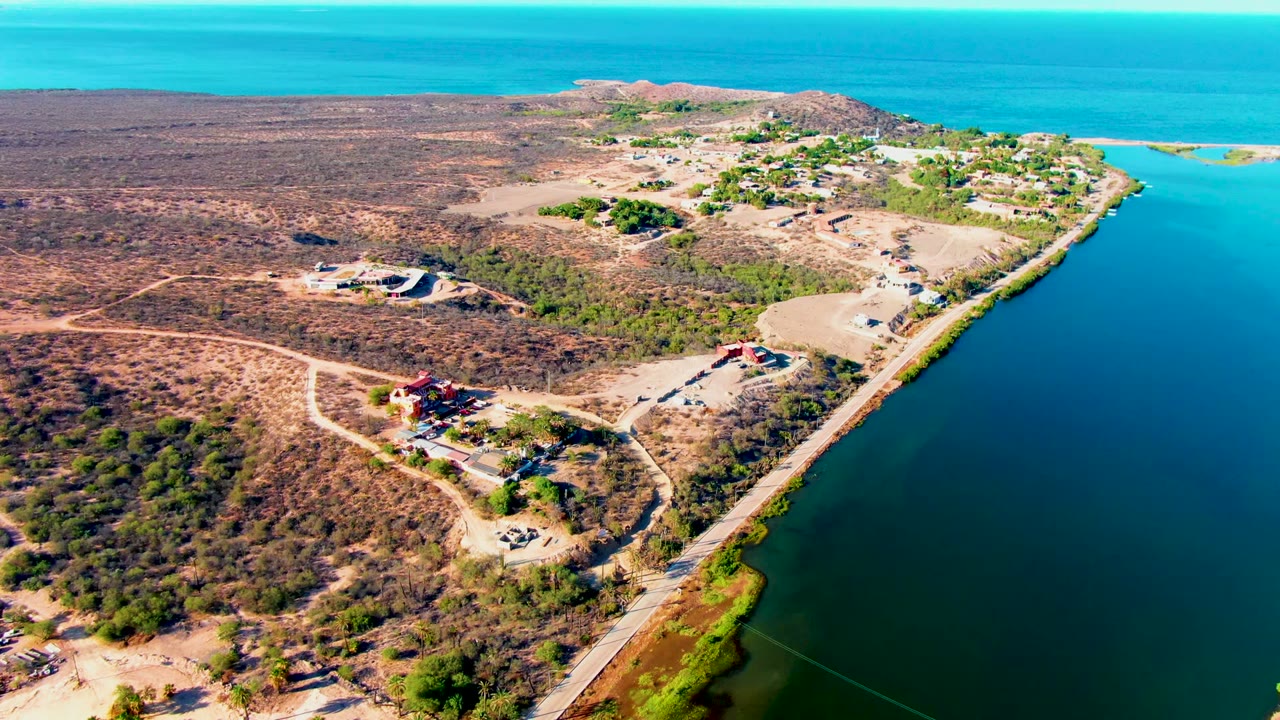 Aerial frame over Mulege, Baja California Sur showing dry brown terrain, scattered buildings, a shoreline road, blue water to the right, low hills in the distance, and no visible people.