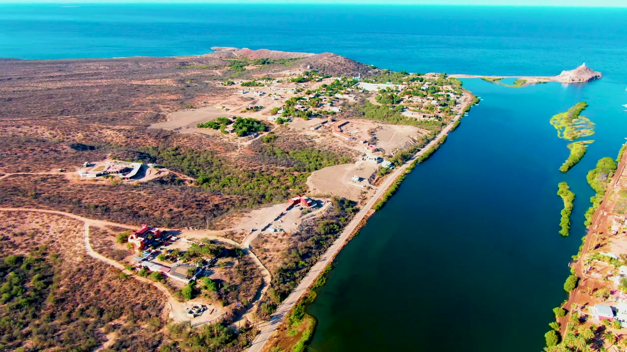 Aerial view in Mulege, Baja California Sur, showing a road beside dark blue water, dry hillside terrain, scattered buildings, and no visible people at the start of the clip.