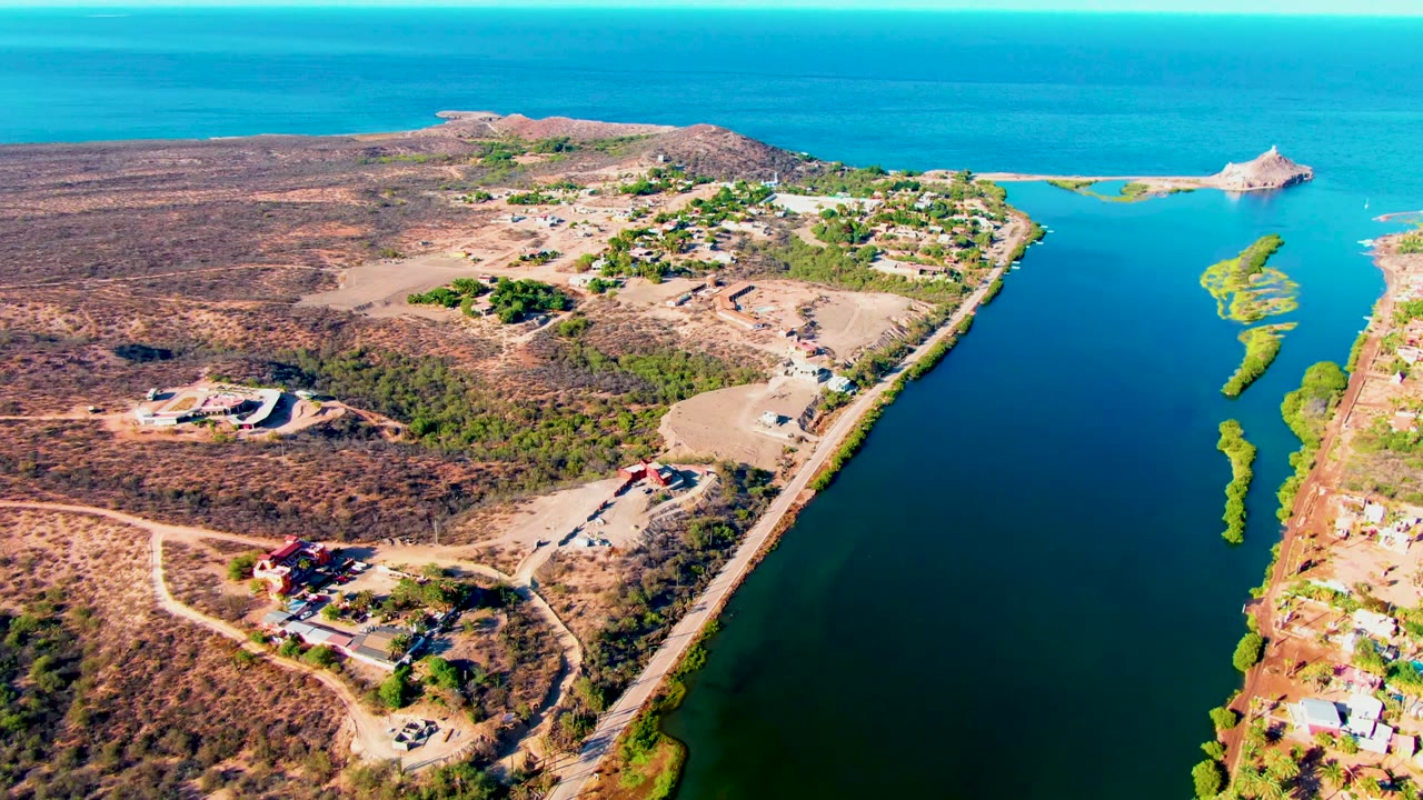 High aerial frame in Mulege, Baja California Sur, showing the road between dry land and a broad water channel, with small buildings, shoreline greenery, and no visible people.