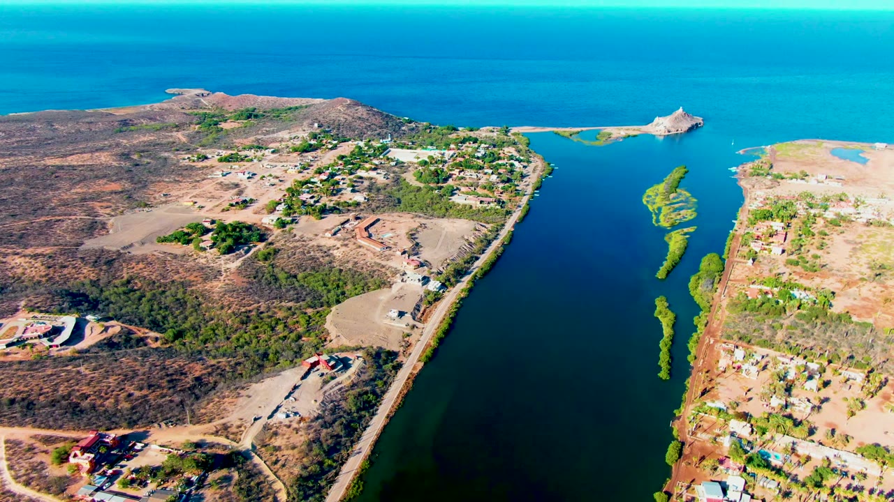 Aerial view over Mulege, Baja California Sur, showing a narrow waterway, coastal road, scattered buildings, dry terrain, turquoise ocean, and no visible people.
