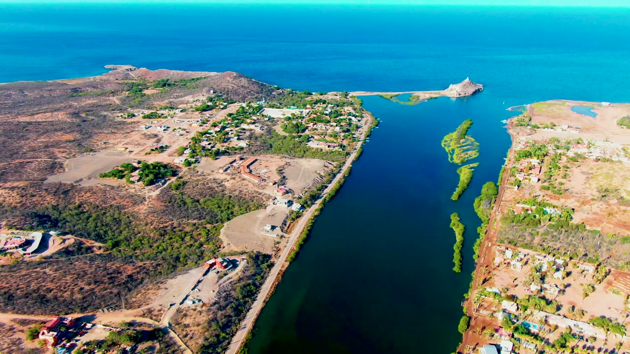 Aerial view in Mulege showing dry coastal terrain on both sides of a narrow waterway, small buildings near the road, ocean horizon, and no visible people.