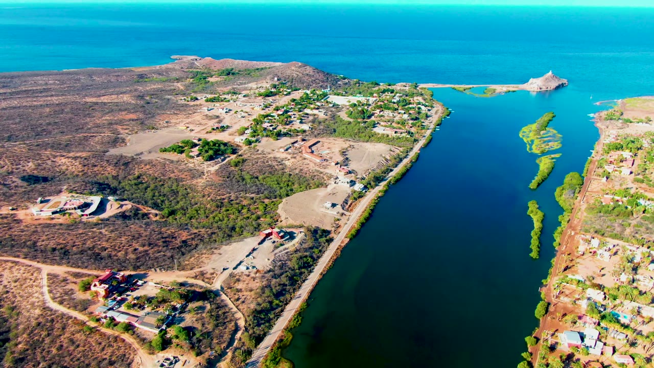 Aerial frame over Mulege, Baja California Sur showing a blue-green canal between dry brown land, scattered buildings, shoreline vegetation, open sea on the horizon, and no visible people.