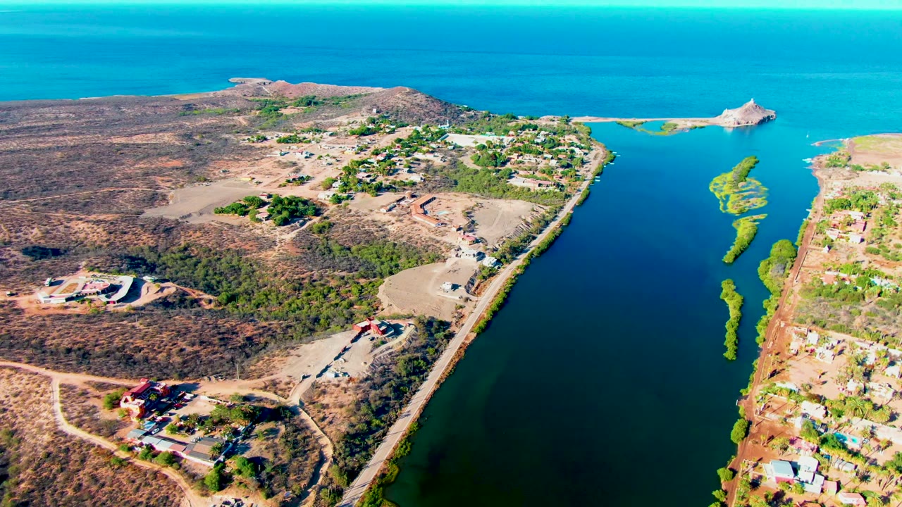 Drone view looking along a canal with dry terrain, homes, and open sea beyond.