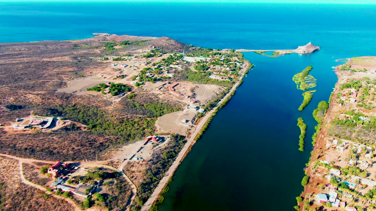 High aerial frame in Mulege showing the canal bordered by brown scrubland and scattered homes, with turquoise sea, a sandy edge, small green islets, and no visible people.