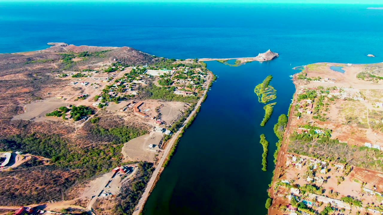 Opening aerial frame over a narrow canal in Mulege, Baja California Sur, with dry land, scattered buildings, green algae in the water, blue ocean beyond, and no visible people.