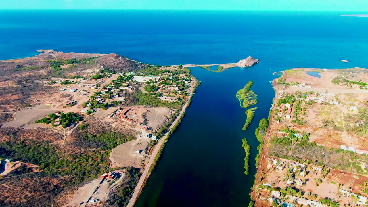 Mid-clip aerial frame over Mulege, Baja California Sur, with deep blue canal water, tan open lots, clustered greenery, scattered structures, ocean horizon, and no visible people.