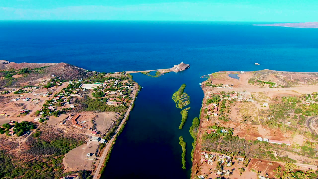 Opening aerial frame over Mulege, Baja California Sur, showing a dark inlet between dry landmasses, scattered buildings, green vegetation, ocean horizon, and no visible people.