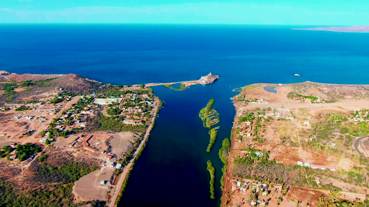 Mid-clip aerial view of Mulege with dark inlet water running toward the ocean, low dry hills and roads on the land, clusters of buildings, green patches, and no visible people.