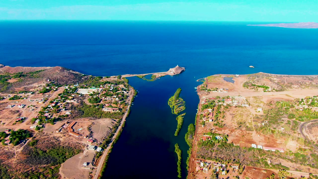 Opening aerial frame over Mulege, Baja California Sur, with a dark inlet between dry landmasses, scattered buildings, turquoise ocean beyond, and no visible people.