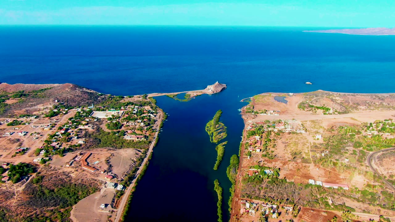 Aerial coastline with inlet, village buildings, dry hills, and distant islands.
