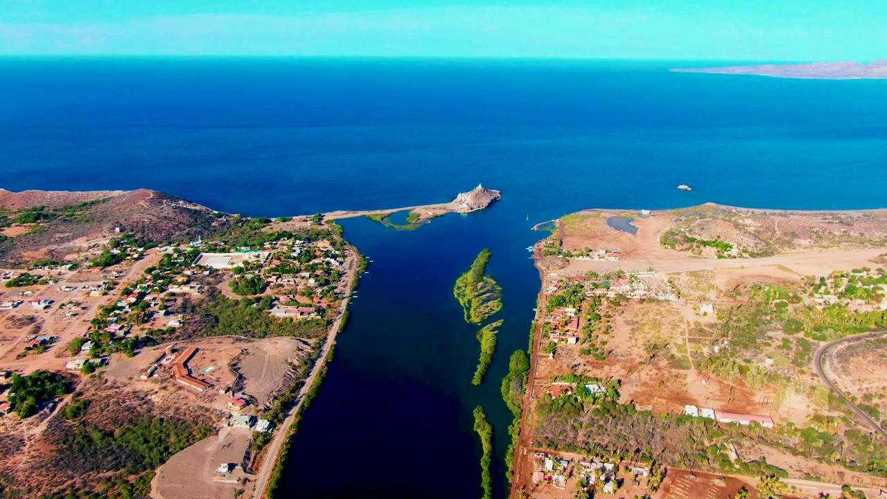 A high aerial view in Mulege shows pale dry terrain on the right, darker inlet water below, a green vegetation strip near the shore, bright ocean beyond, and no visible people.