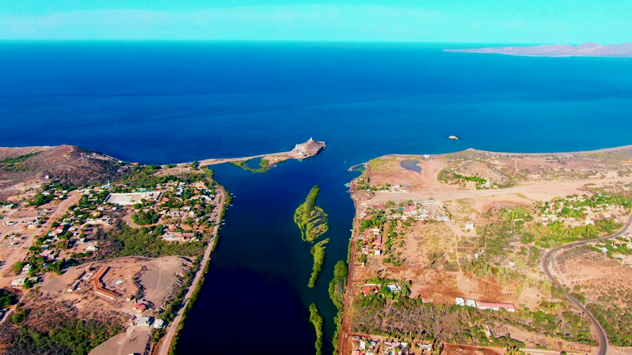 Aerial frame over Mulege, Baja California Sur showing a narrow coastal inlet, dry shoreline, scattered buildings, roads, blue ocean, and no visible people.