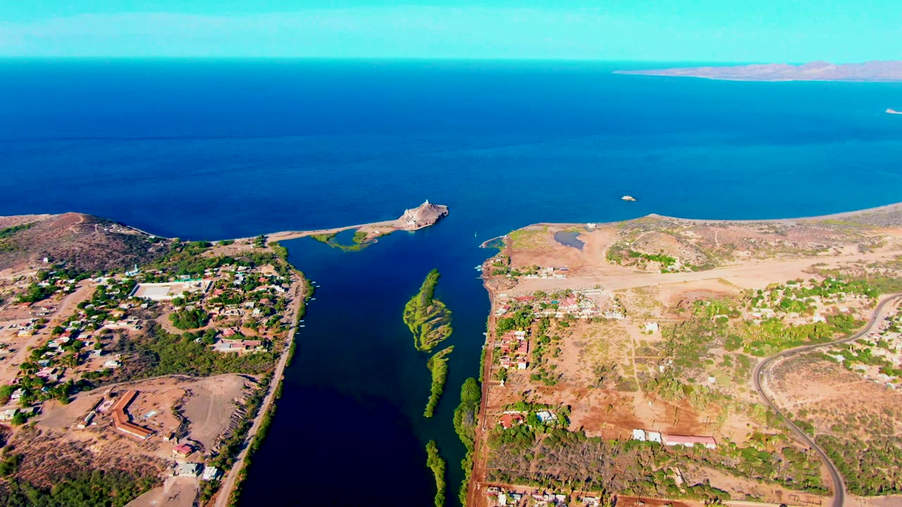 Mulege, Baja California Sur aerial frame showing a small white boat on the blue water near the inlet, with dry shoreline, buildings, roads, and no visible people.
