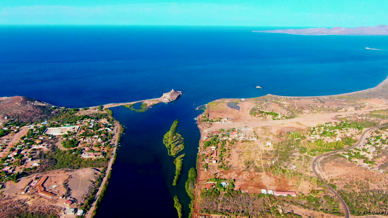 Aerial frame over Mulege, Baja California Sur showing a blue coastal channel, narrow green island, arid shoreline, scattered development, distant ocean, and no visible people.