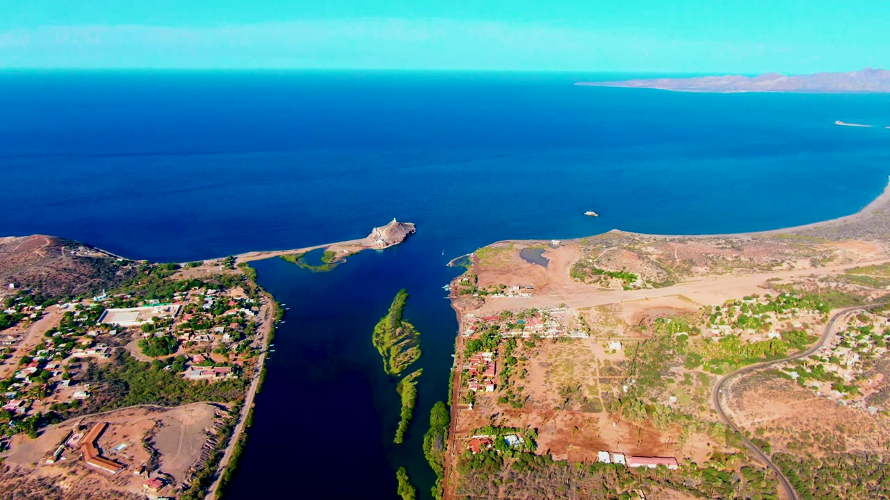 Mulege aerial frame showing the coastal channel, green island strip, arid tan land, scattered structures, a small boat on blue water, distant ocean, and no visible people.