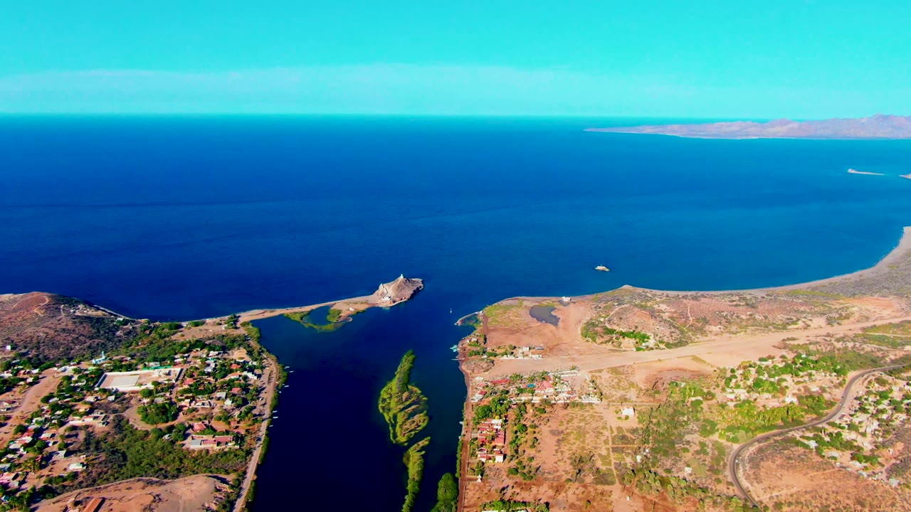 At 0:00, an aerial view over Mulege, Baja California Sur shows dry terrain, scattered buildings, dark inlet water, deep blue ocean, distant land on the horizon, and no visible people.