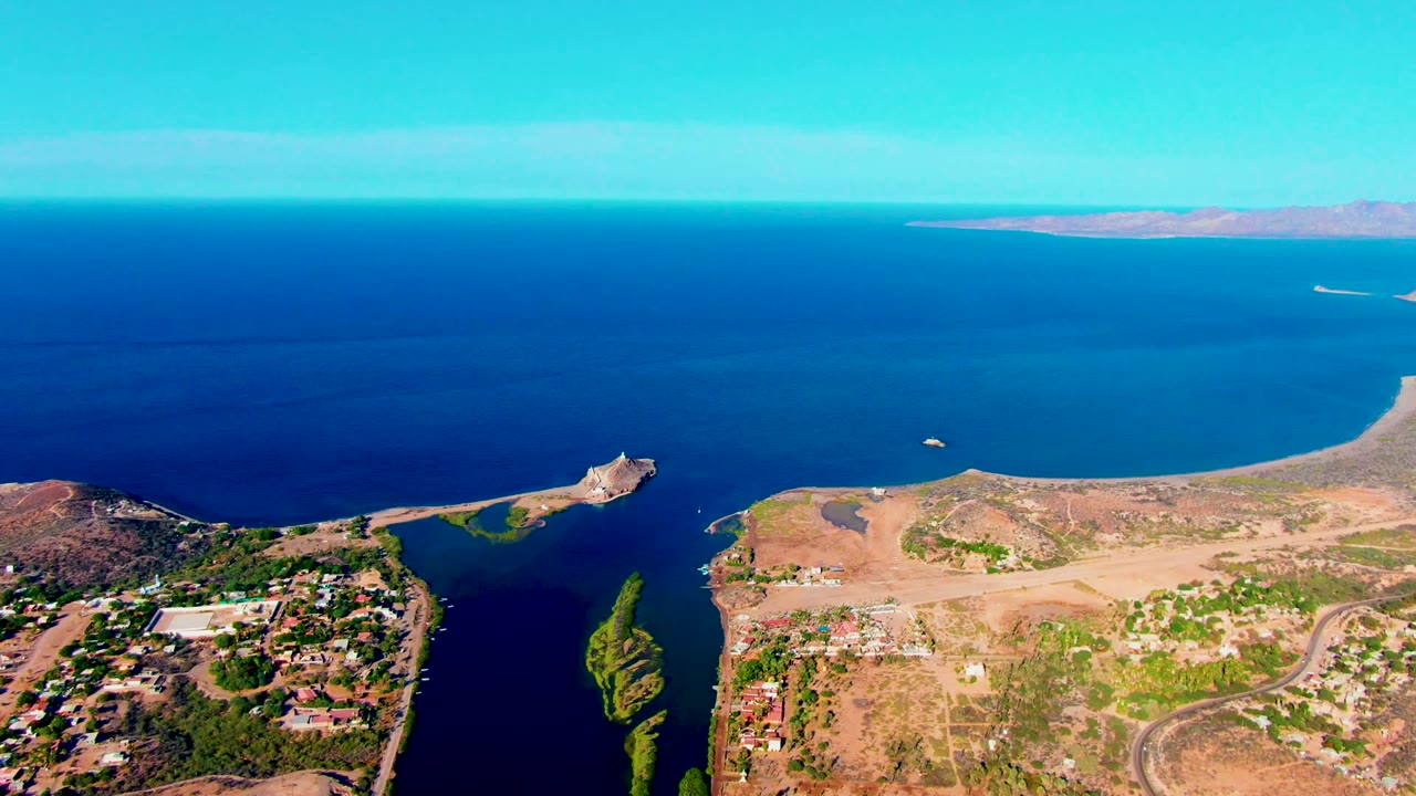 At 0:02, a high aerial frame in Mulege shows green vegetation along dark water channels, dry open ground, scattered structures, broad blue sea, a small white object on the water, and no visible people.