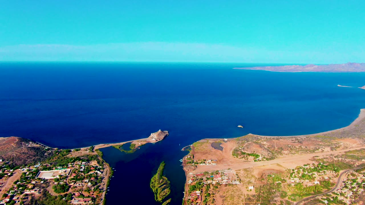Wide aerial view over Mulege, Baja California Sur, with deep blue ocean, a sandy peninsula, scattered buildings at the lower edge, distant land on the horizon, and no visible people.