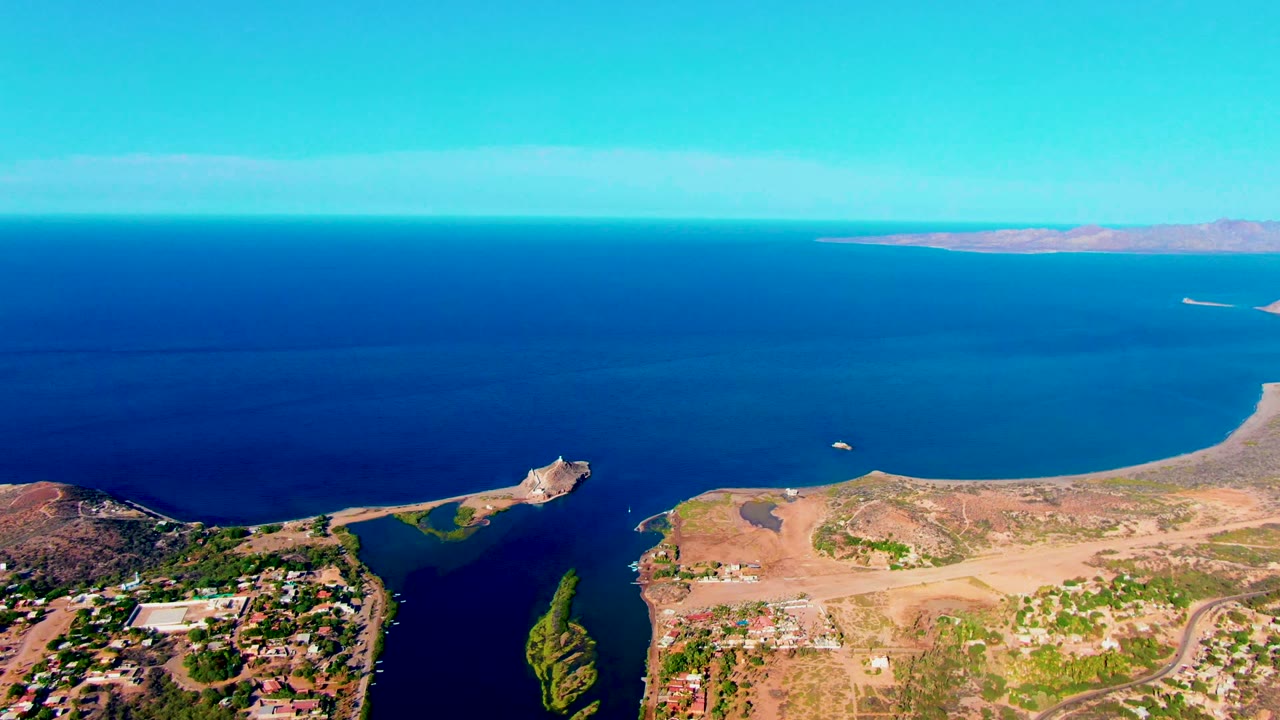 High coastal aerial with ocean, peninsula, buildings, and distant horizon land.