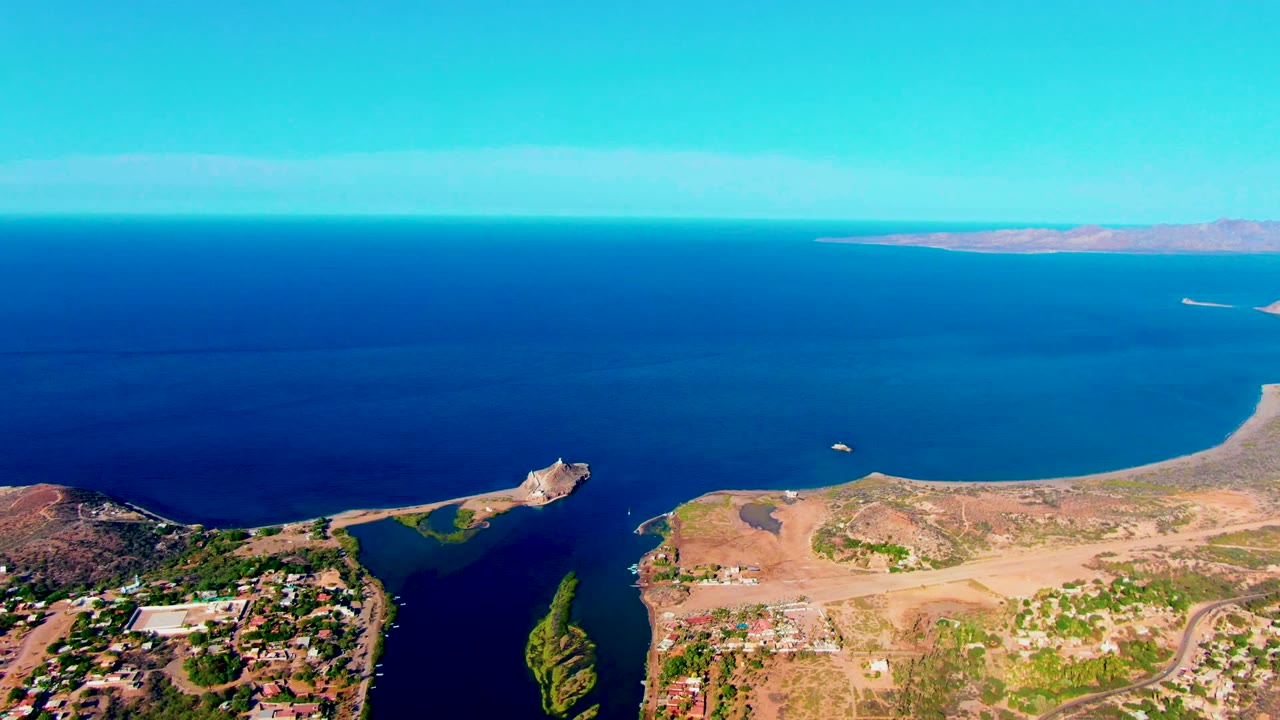 Drone-style aerial over Mulege coastline with a small bright vessel on the water, a curved peninsula below, developed shoreline at the bottom, and no visible people.