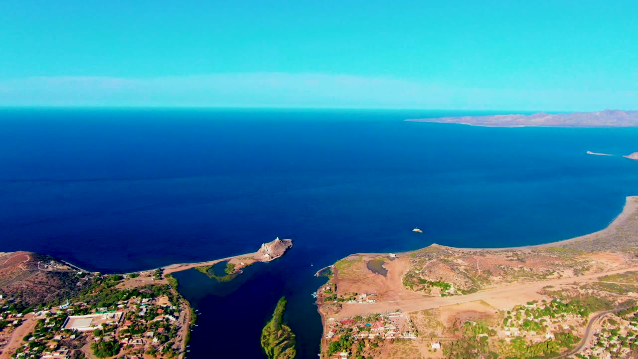 Mulege coastal aerial frame shows deep blue water filling most of the view, tan peninsula land below, distant islands on the horizon, scattered buildings, and no visible people.
