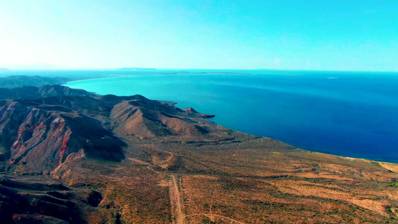 Opening aerial frame over Mulege, Baja California Sur, showing dry mountain ridges, scrub terrain, blue ocean, clear sky, and no visible people.