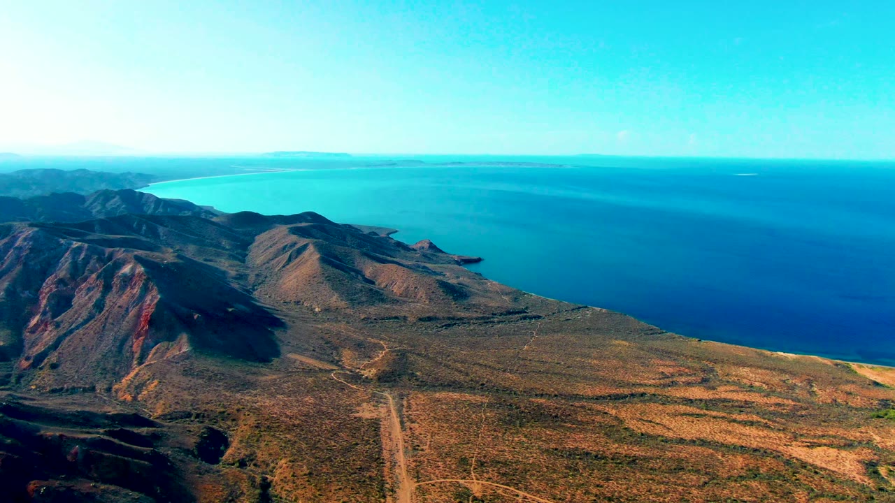 Wide aerial coastline with rocky hills, dry terrain, and ocean horizon.